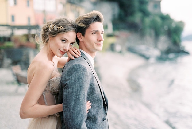 La novia abraza al novio por detrás en la orilla del lago