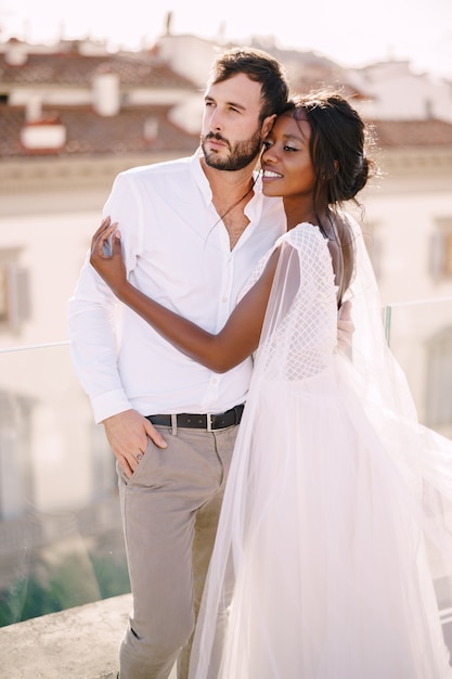 Novia afroamericana abraza al novio caucásico en el techo con vista al paisaje urbano