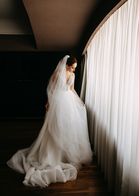 Novia ajustando su vestido, de pie a la luz de la ventana.