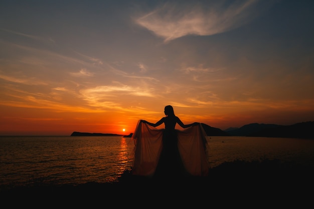 la novia al atardecer se para en la orilla del mar y sostiene los extremos del vestido en su estirado