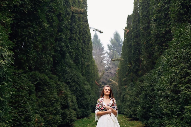 La novia se para entre los árboles en el parque con vestido de novia y un chal