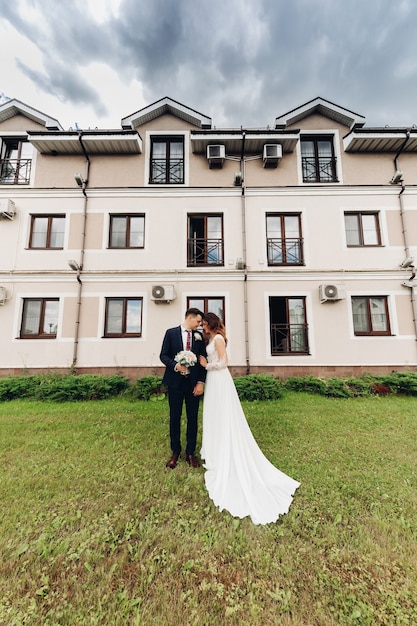 Novia atractiva con vestido blanco largo ama a su fuerte marido cerca de la hermosa construcción