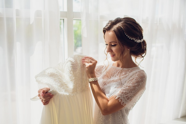 Novia bastante morena en bata de seda y velo de encaje con vestido de novia en una percha cerca de la ventana.