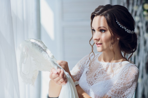 Foto novia bastante morena en bata de seda y velo de encaje con vestido de novia en una percha cerca de la ventana.