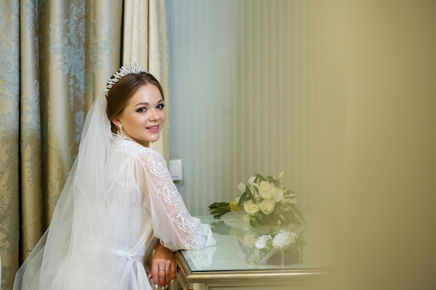 La novia con una bata blanca y un velo se sienta en la mesa de maquillaje