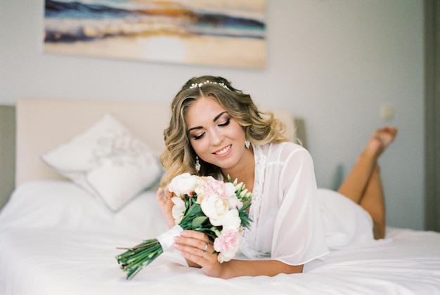 La novia con una bata blanca yace boca abajo en la cama mirando un ramo de flores