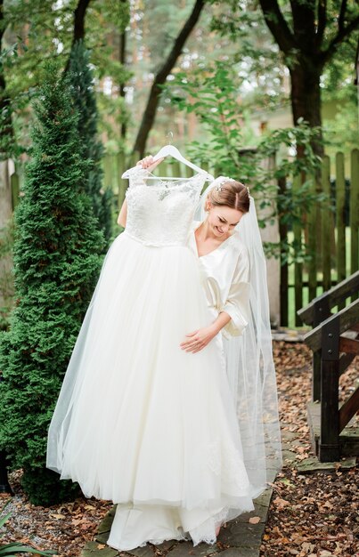Novia en bata de seda trata de vestidos de novia de pie en el porche cubierto con hojas caídas
