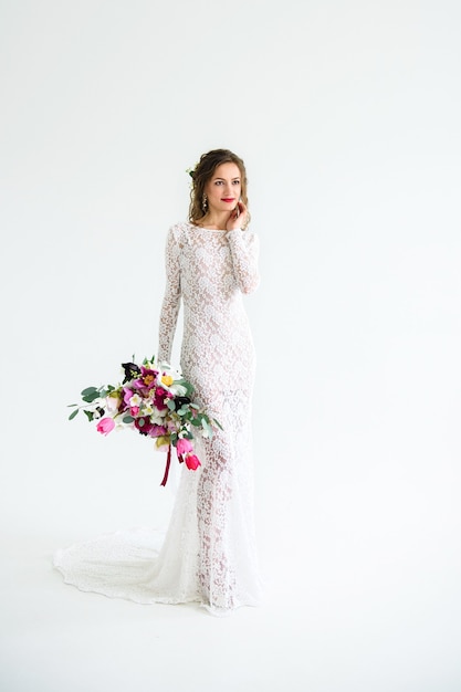 Novia chica alegre en un vestido de punto blanco posando con un ramo de flores