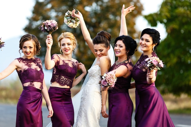 La novia y las damas de honor en vestidos violetas bailan en el camino