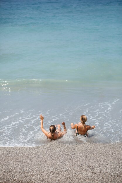 Novia de dos niñas están sentadas en la orilla arenosa del mar y las olas las empaparon en trajes de baño en un día cálido y soleado