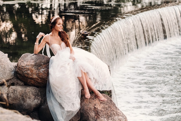 Una novia elegante con un vestido blanco, guantes y los pies descalzos está sentada cerca de una cascada en el parque disfrutando de la naturaleza.Una modelo con un vestido de novia y guantes en un parque natural.Belarús