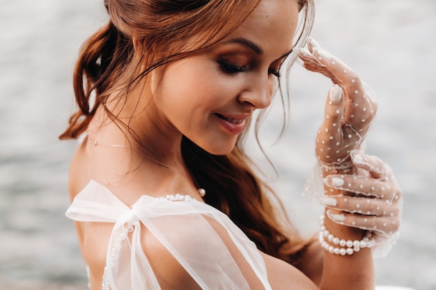 Una novia elegante con un vestido blanco, guantes y los pies descalzos está sentada cerca de una cascada en el parque disfrutando de la naturaleza.Una modelo con un vestido de novia y guantes en un parque natural.Belarús