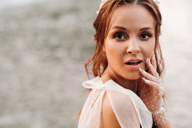 Una novia elegante con un vestido blanco, guantes y los pies descalzos está sentada cerca de una cascada en el parque disfrutando de la naturaleza.Una modelo con un vestido de novia y guantes en un parque natural.Belarús