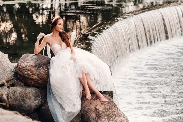 Una novia elegante con un vestido blanco, guantes y los pies descalzos está sentada cerca de una cascada en el parque disfrutando de la naturaleza.