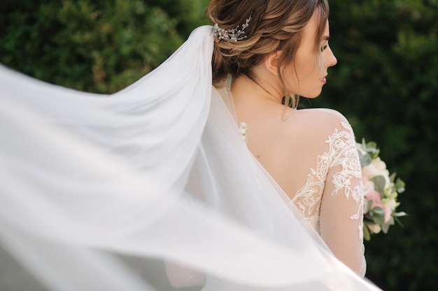 Novia elegante en vestido de novia blanco sostiene ramo de flores. Gran viel. Fondo de arbusto verde