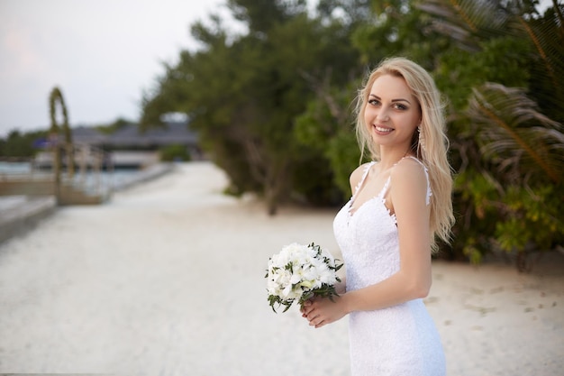 La novia feliz con un ramo de flores blancas en la playa del balneario tropical de lujo está lista para la ceremonia de la boda