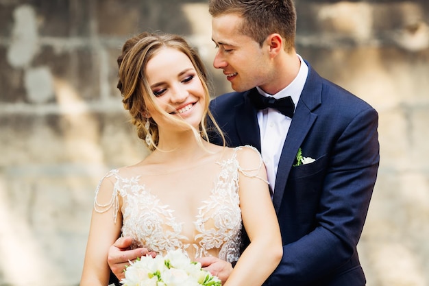 La novia feliz con un vestido blanco con un hermoso maquillaje sostiene un ramo de novia y un novio que está a su lado