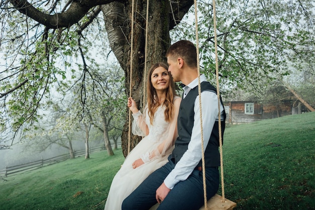 La novia con la figura perfecta se sienta en el columpio, el esposo se sienta al lado. Disparo al aire libre, parque de primavera o concepto de verano. Amor, gente sensual