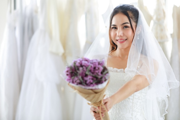 Novia hermosa asiática en vestido blanco con velo de encaje de pelo transparente de pie sonriendo mirar a cámara con ramo de flores de boda fragante púrpura en las manos en primer plano borroso en el vestidor.