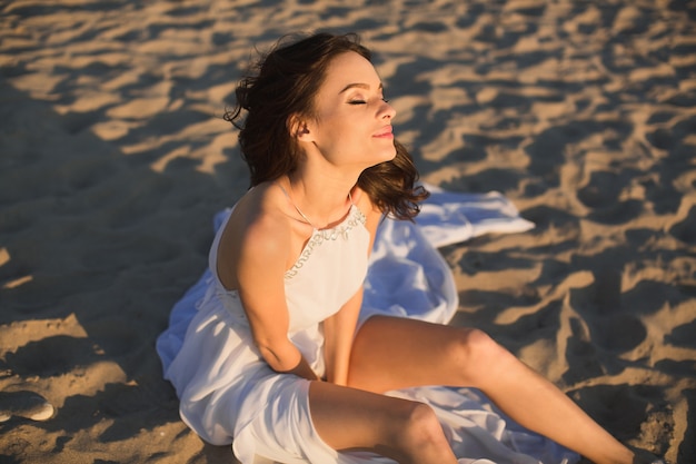 Novia hermosa chica con un vestido blanco sentada en la arena de la playa al atardecer