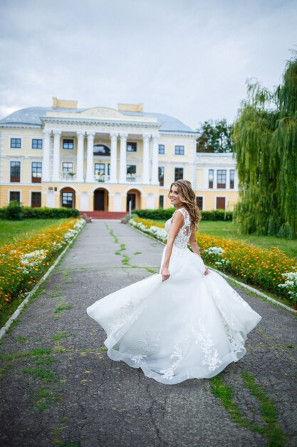 Novia hermosa chica con un vestido blanco con un tren y un paseo en el contexto de una gran casa con columnas en el día de su boda