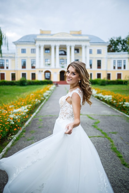 Novia hermosa chica con un vestido blanco con un tren y un paseo en el contexto de una gran casa con columnas en el día de su boda