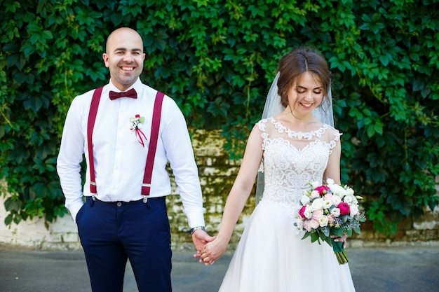 Novia hermosa pareja en un vestido blanco con un ramo de flores mientras el novio con tirantes y pajarita cogidos de la mano. En el contexto de una pared con hojas verdes. Pareja feliz. Concepto de matrimonio.