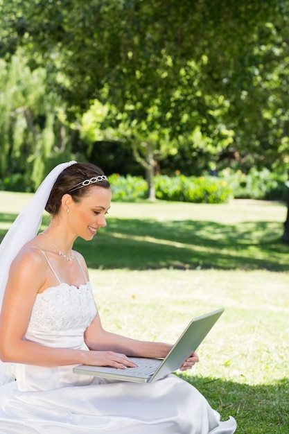 Novia hermosa que usa la computadora portátil mientras que se sienta en hierba en el jardín