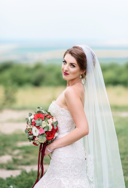 La novia hermosa en el vestido blanco con clase presenta con el ramo rojo de la boda en un césped verde