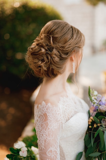 Una novia con un hermoso peinado con un vestido de encaje se encuentra con un ramo y mira hacia un lado