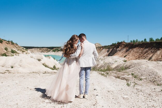 La novia en un hermoso vestido abrazando al novio en un traje ligero cerca del lago. Novios de pie en una colina de arena al aire libre. Una romántica historia de amor. Agua azul celeste en el horizonte.