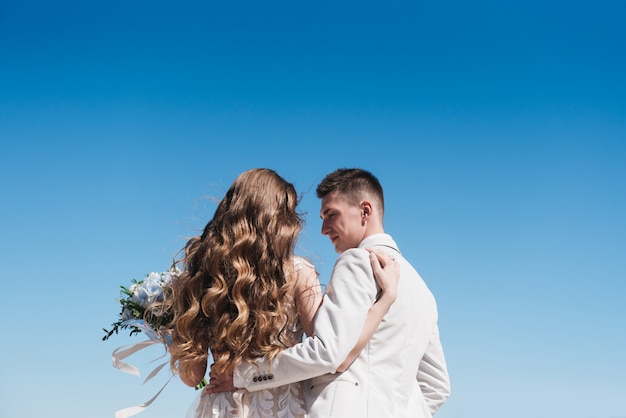 La novia en un hermoso vestido abrazando al novio en un traje ligero contra el cielo azul. Una romántica historia de amor.