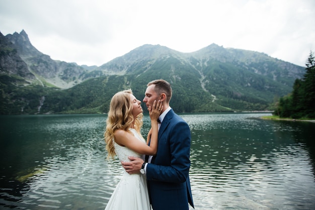 Novia con hermoso vestido blanco y novia con vistas a hermosas montañas verdes y lago con agua azul