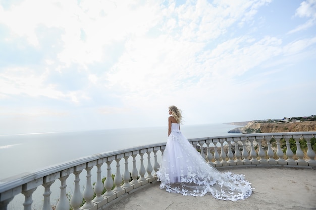 Una novia con un hermoso vestido de novia blanco se encuentra en un balcón en la cima de una montaña en un soleado día de verano