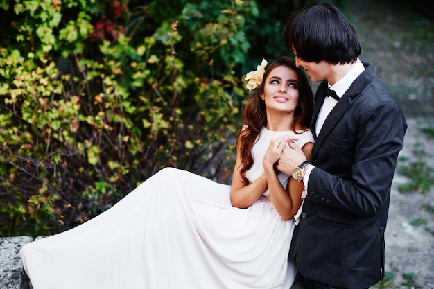 Novia increíble con el pelo largo y rizado y el novio de pie cerca uno del otro en hojas verdes, foto de boda, hermosa pareja.