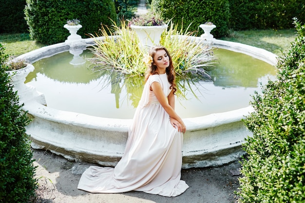 Novia increíble con el pelo largo y rizado en vestido de novia sentado junto a la fuente en el fondo del parque, foto de boda, retrato.