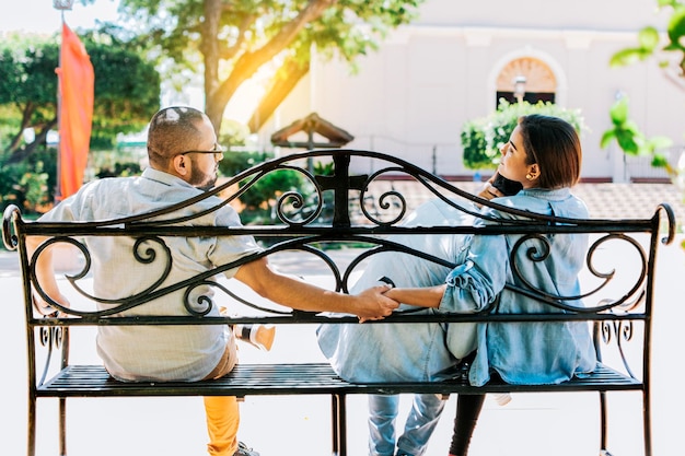 Novia infiel con su novio sosteniendo la mano de otro hombre en un parque Mujer infiel sentada de la mano con otro hombre mientras su novio abraza su concepto de triángulo amoroso