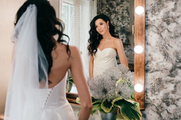 Novia joven hermosa en el vestido de boda blanco que mira en espejo.