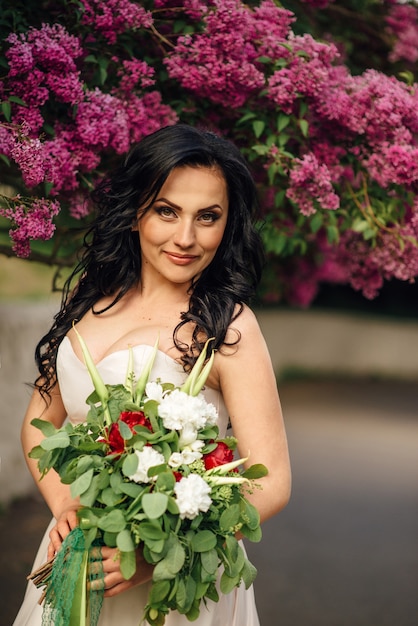 Novia joven con un lujoso vestido de flores está de pie en un jardín lila floreciente