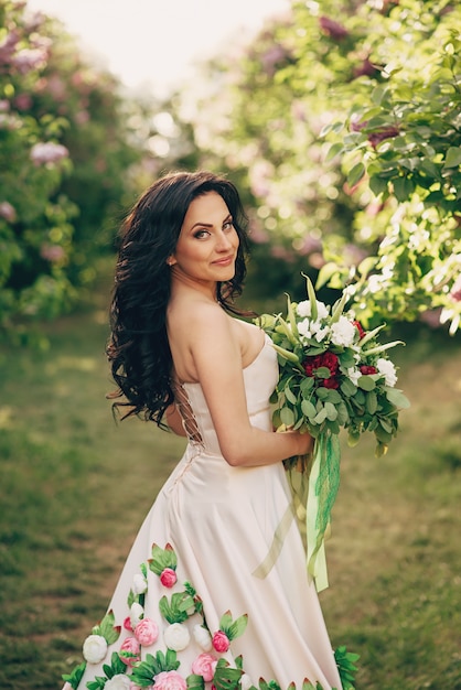 Novia joven con un lujoso vestido de flores está de pie en un jardín lila floreciente