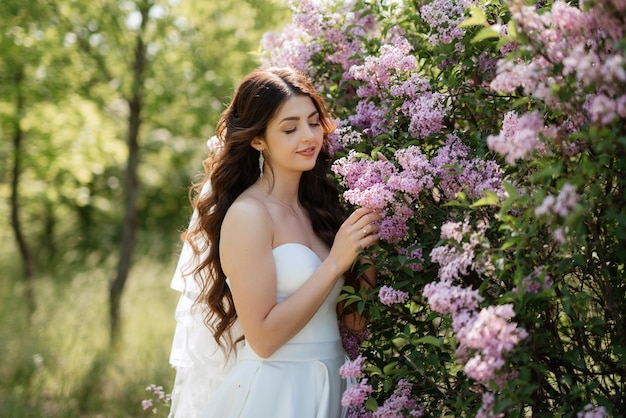 Novia joven con un vestido blanco en un bosque de primavera en arbustos de lilas