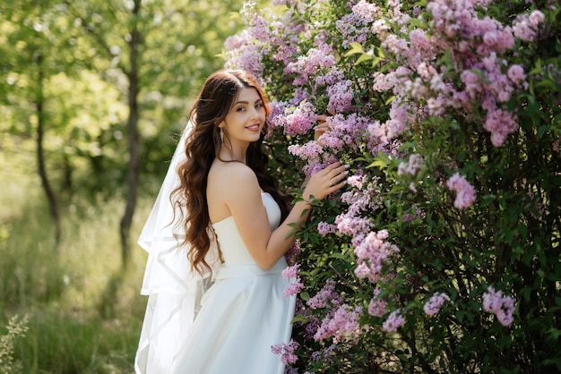 Novia joven con un vestido blanco en un bosque de primavera en arbustos de lilas