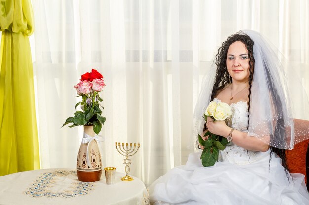 Una novia judía feliz con un ramo de rosas blancas se sienta en la sinagoga antes de la ceremonia de Huppa en una mesa con flores y una menorá. Foto horizontal.