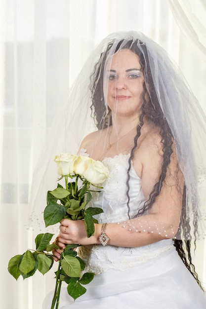 Novia judía con el rostro cubierto con velo antes de una ceremonia de chuppa con un ramo de rosas blancas en sus manos. Foto vertical