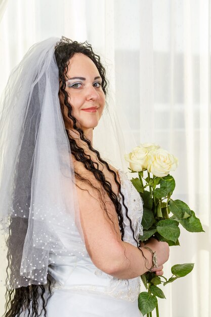 Una novia judía se para en el salón antes de la ceremonia de la chuppa con un ramo de rosas blancas en sus manos, una foto en la cintura. Foto vertical