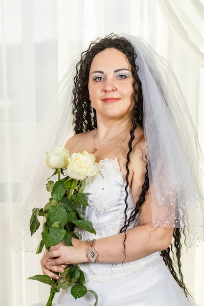 Una novia judía se para en el salón antes de la ceremonia de la chuppa con un ramo de rosas blancas en sus manos, una foto en la cintura. Foto vertical