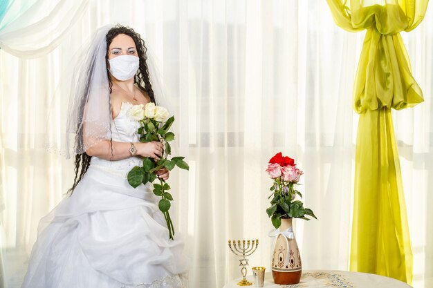 Una novia judía en una sinagoga en una mesa con flores antes de una ceremonia de chuppa durante una pandemia, con una máscara médica y un ramo de flores, espera al novio