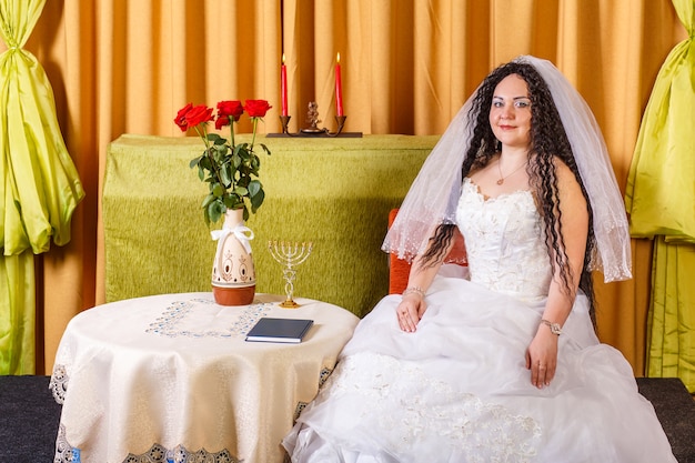 Una novia judía con un vestido de novia blanco con velo se sienta en una mesa con flores antes de la ceremonia de la jupá. Foto horizontal