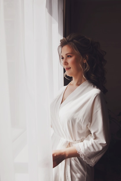 Novia linda joven en albornoz de casa en la ventana en la habitación del hotel. Mañana de la novia el día de la boda. Feliz mujer está esperando para conocer a su novio. Concepto de matrimonio feliz y de lujo. Copia espacio