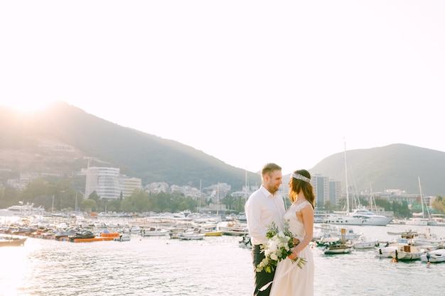 La novia y el novio se abrazan y miran en el contexto del muelle de barcos en budva